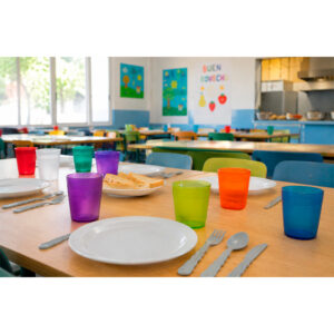 Niños sentados en un comedor escolar compartiendo mesa con jarras de agua, vasos apilables y platos